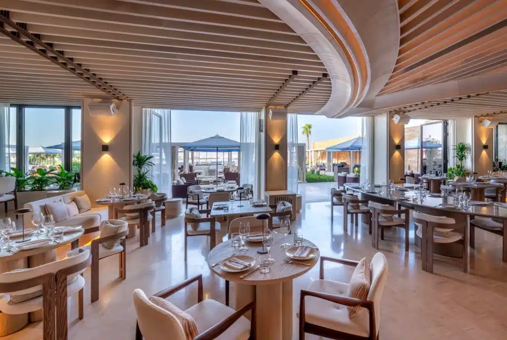 Stylish Ilios Beach Club restaurant interior with wooden ceiling, round tables set for dining, and large windows overlooking a patio with umbrellas.