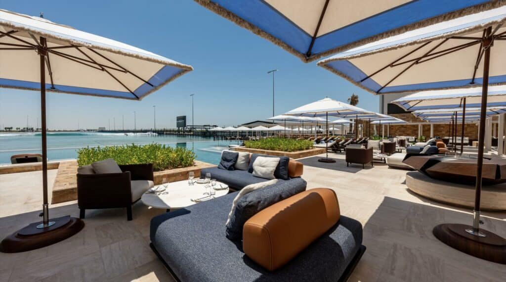Outdoor lounge area with plush seating, white umbrellas, and a view of the water under a clear blue sky.