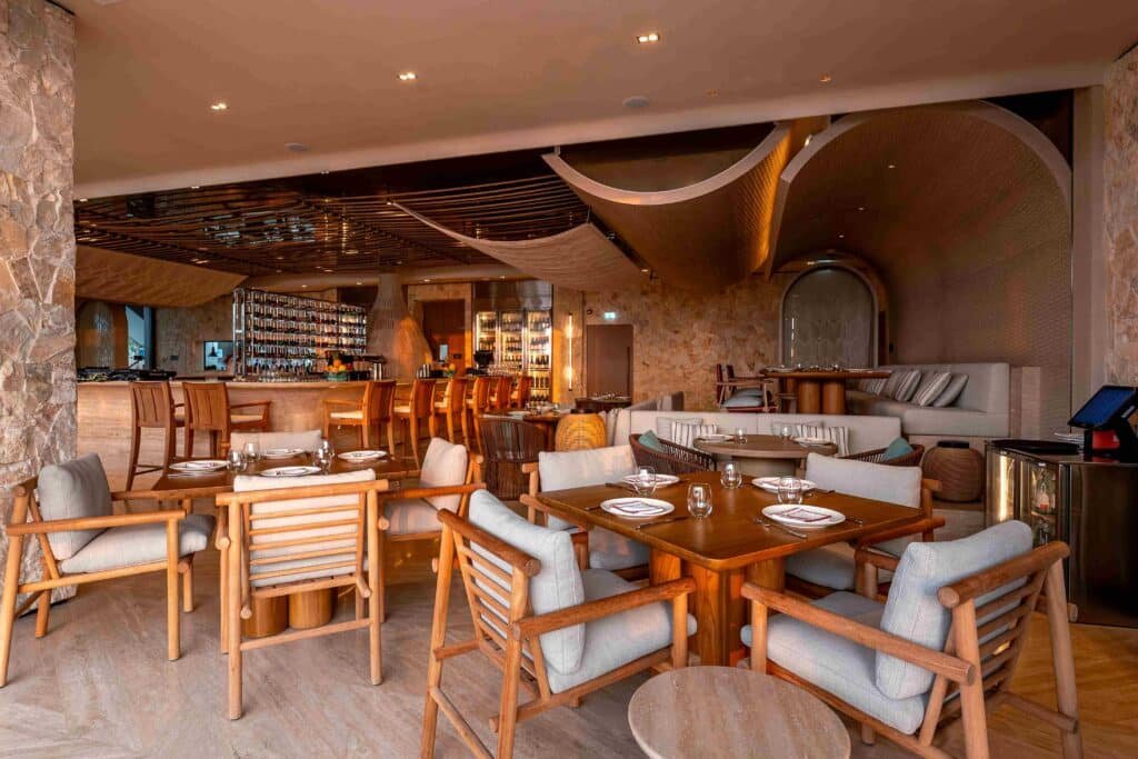 A modern restaurant interior with wooden furniture and stone accents, featuring a bar area with high stools and neatly set tables.