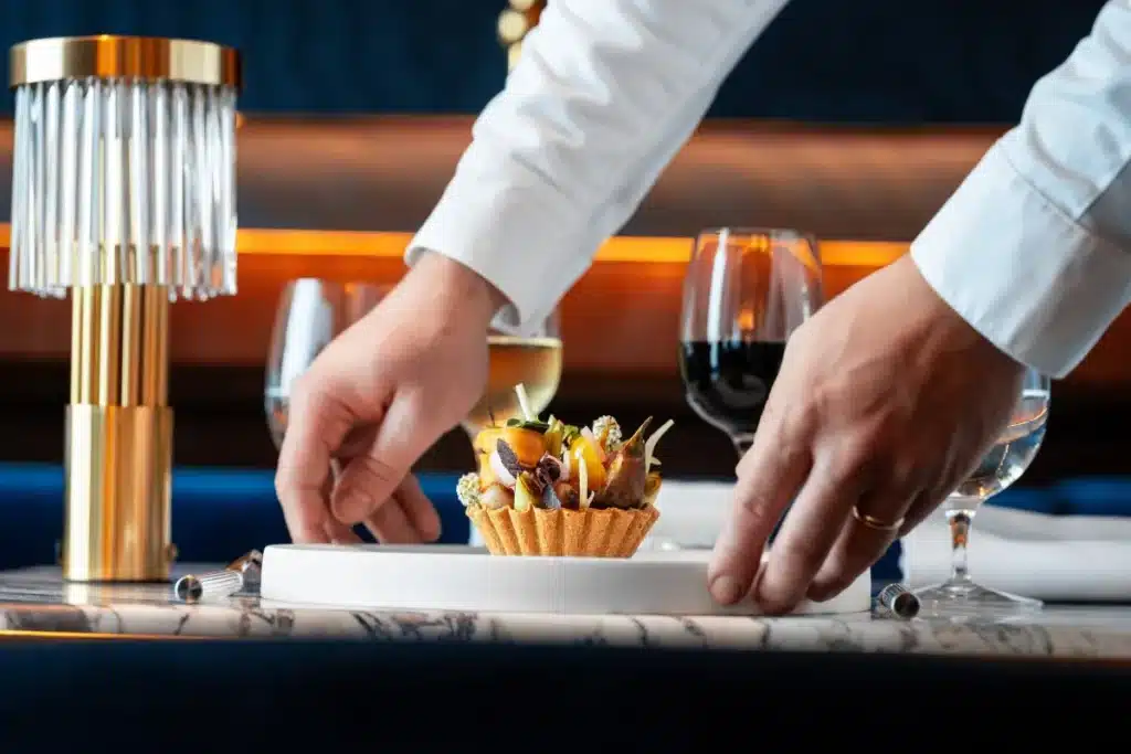 A server in a white shirt places a dessert tart on a table set with wine glasses and a modern lamp nearby.