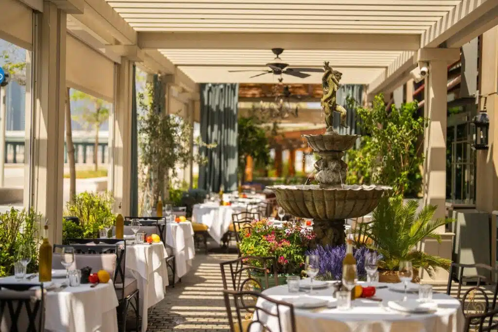 Sunlit outdoor restaurant patio with white tablecloths, a central fountain, and plants under a pergola