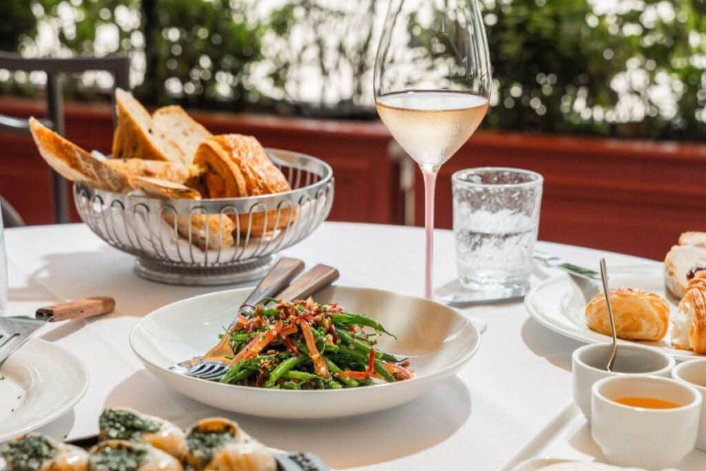 Table with a dish of vegetables and chopsticks, a basket of bread, a glass of wine, and various side dishes.