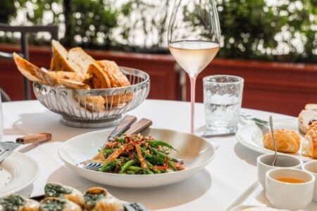 Table with a dish of vegetables and chopsticks, a basket of bread, a glass of wine, and various side dishes.