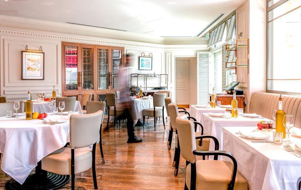 Elegant La Vie En Rose Brunch restaurant interior with neatly set tables, white tablecloths, and bottles of olive oil; a blurred figure moves between tables.