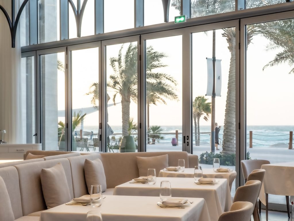 Upscale beachfront restaurant with set tables, beige seating, and large glass doors overlooking palm trees and the ocean