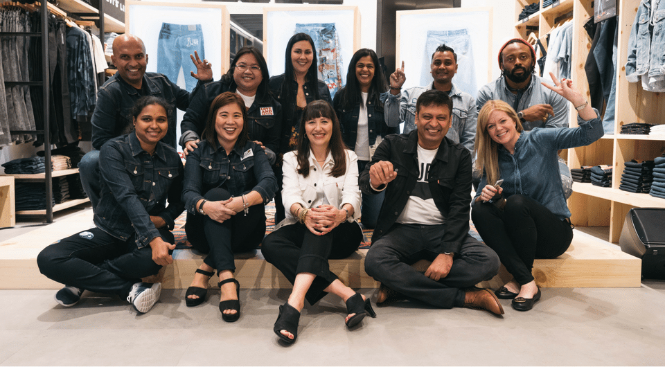 A group of smiling people wearing denim jackets sitting and standing in front of displayed Levi's 501 Day jeans within a clothing store.