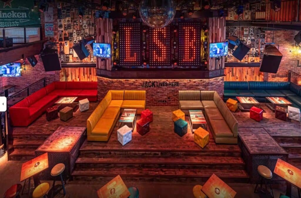 Colorful nightclub lounge with couches, cube stools, and a stage lit by an LED sign reading LSBI