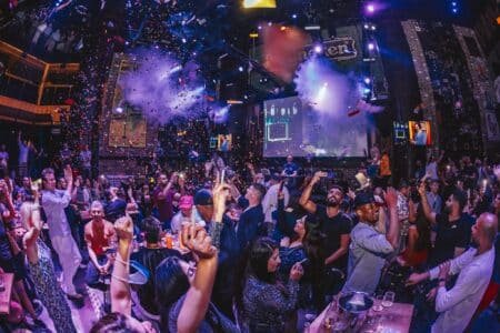 Crowded nightclub with confetti, smoke, and people cheering under colorful stage lights