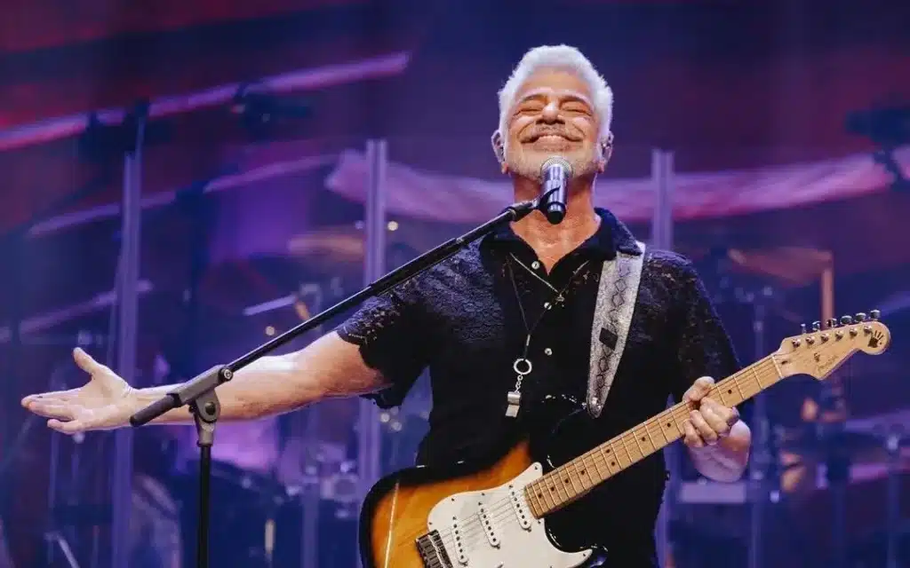 A man with white hair, wearing a black shirt, playing an electric guitar and smiling on stage.