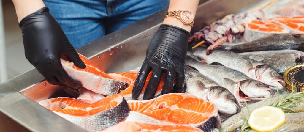 A person wearing black gloves arranges salmon fillets on ice next to whole fish and lemon slices in a seafood market.