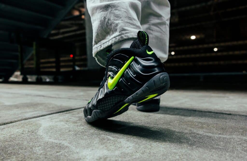 A person wearing black sneakers with neon green accents and a prominent swoosh design walks on a concrete surface.