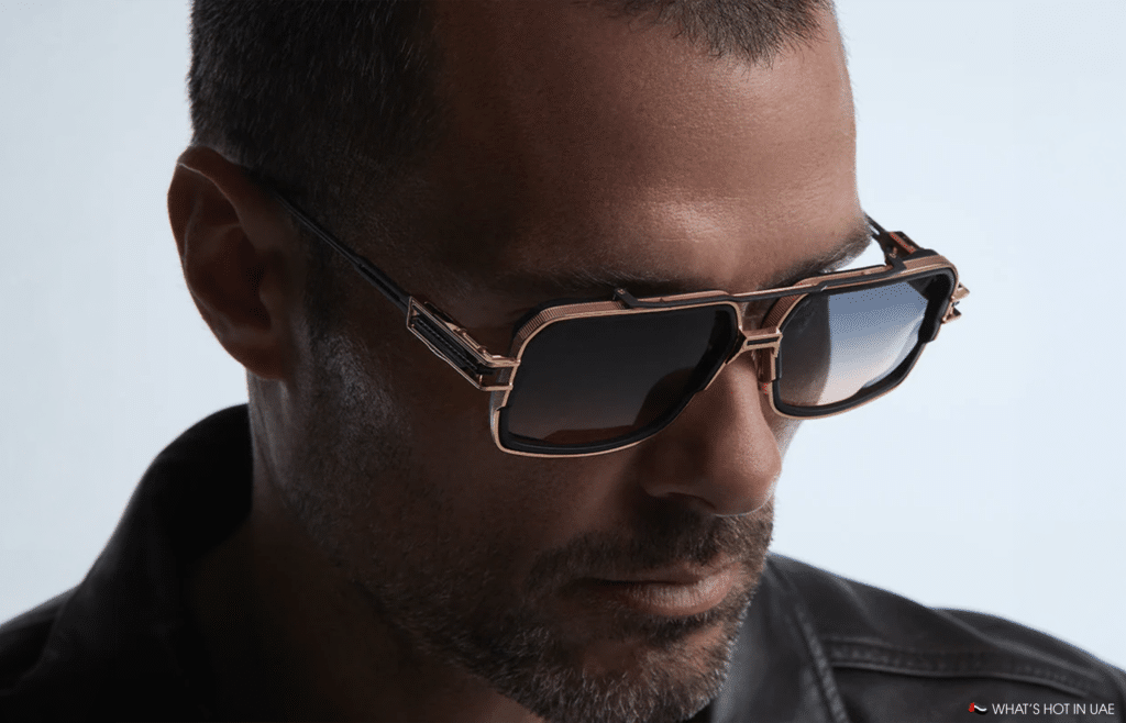 Close-up of a man wearing gold-framed sunglasses and a dark jacket against a light background