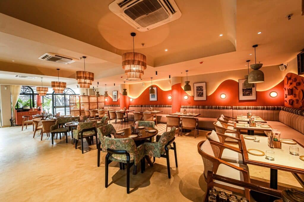A spacious, warmly lit Pincode By Kunal Kapur restaurant interior with neatly arranged tables, patterned chairs, and decorative lighting fixtures.