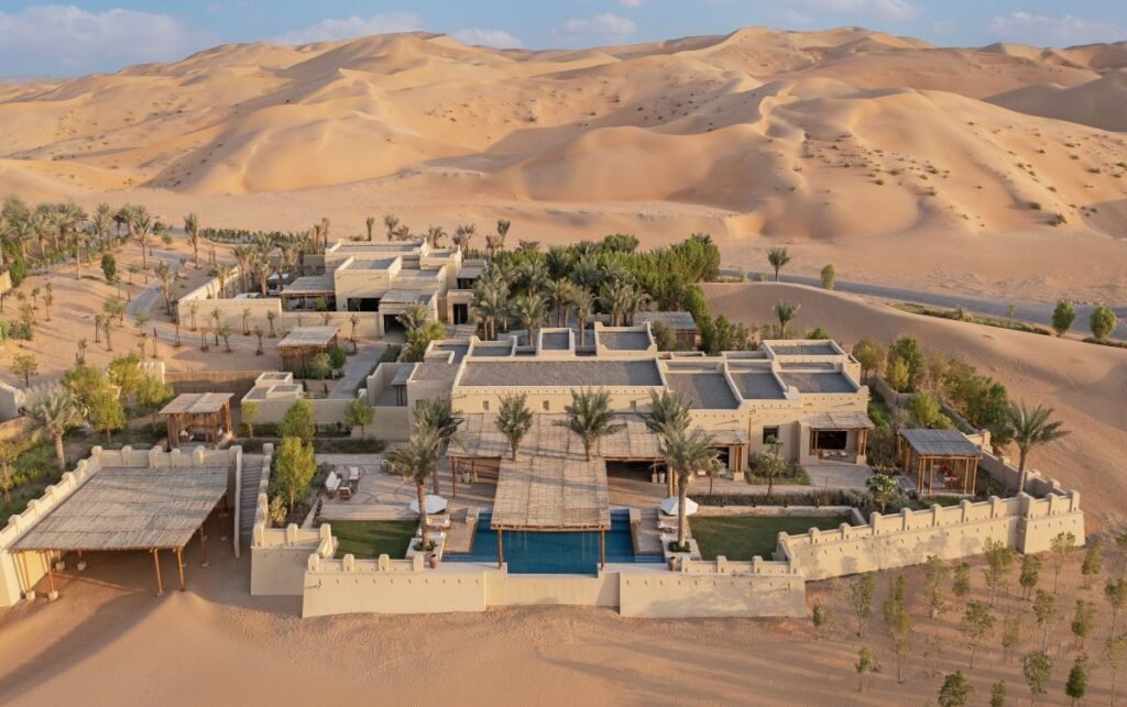 A luxurious desert resort surrounded by sand dunes, featuring traditional architecture, a pool, and lush greenery.