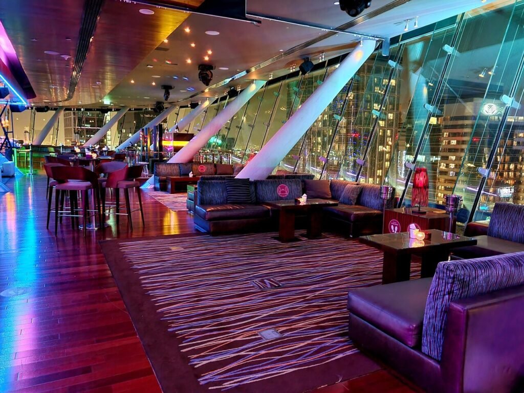 Modern rooftop lounge with couches, tables, and colorful city lights seen through glass walls at night