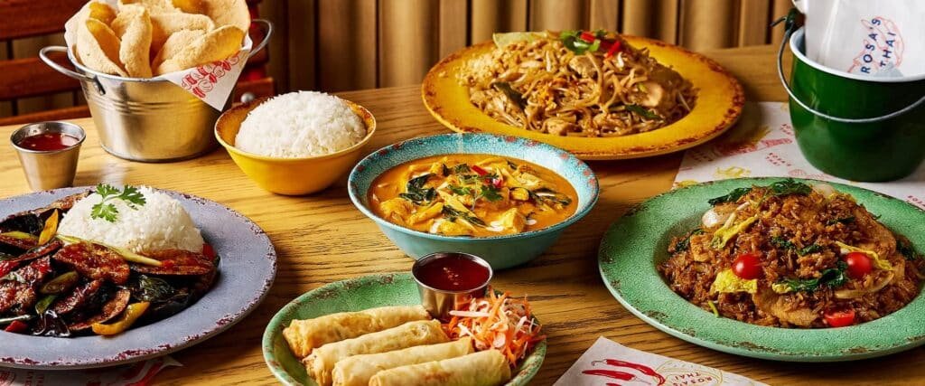 A spread of various Asian dishes including rice, curry, spring rolls, and stir-fried noodles on a wooden table.