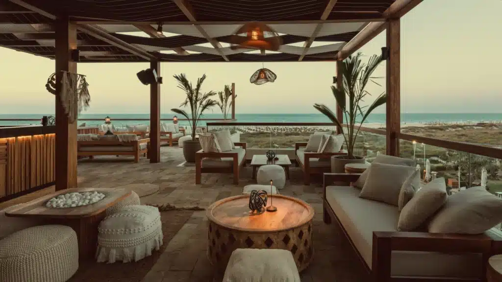 Outdoor patio with wooden furniture, cushions, palm plants, and views of the ocean under a pergola at sunset.