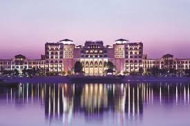 A grand illuminated hotel or palace reflected in calm water at dusk under a purple sky