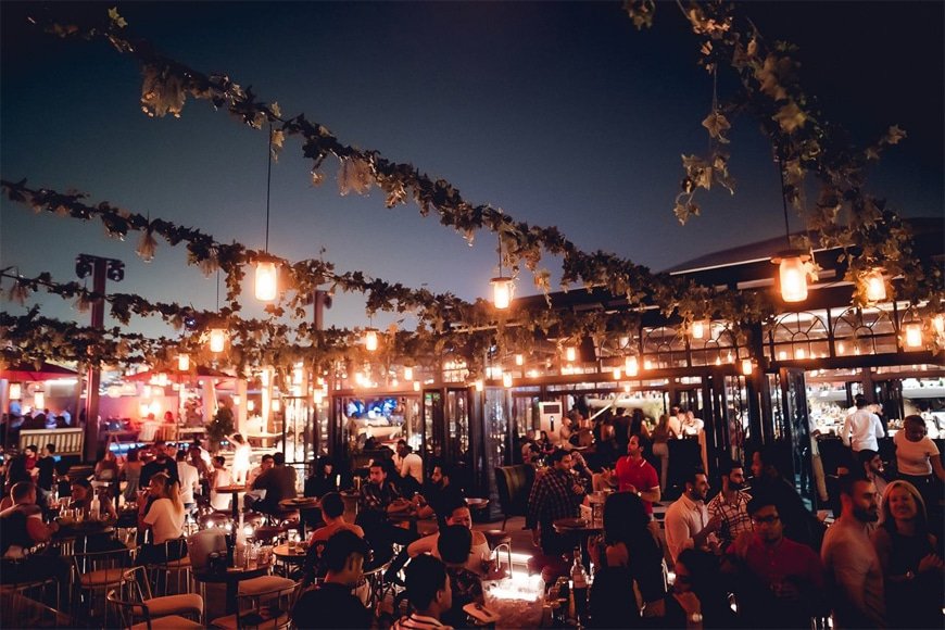 Crowded outdoor rooftop restaurant at night with hanging lights and vine-covered pergolas