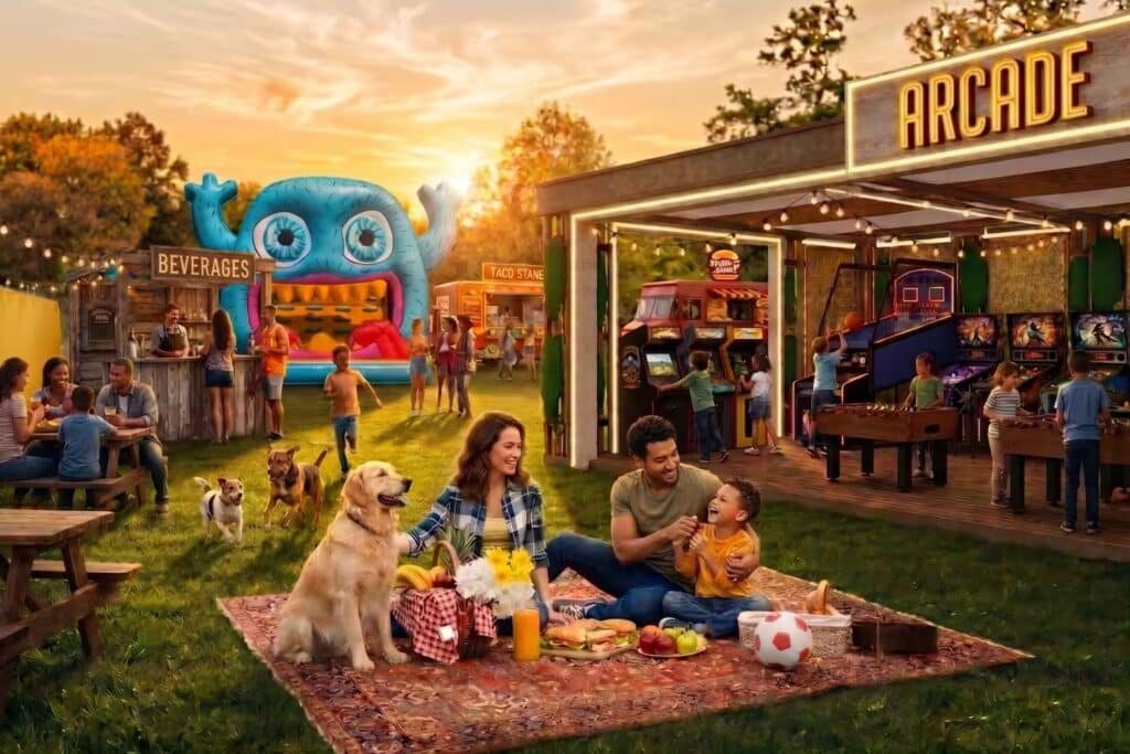 A family having a picnic on a lawn near an arcade and inflatable play area, with people dining and dogs nearby.