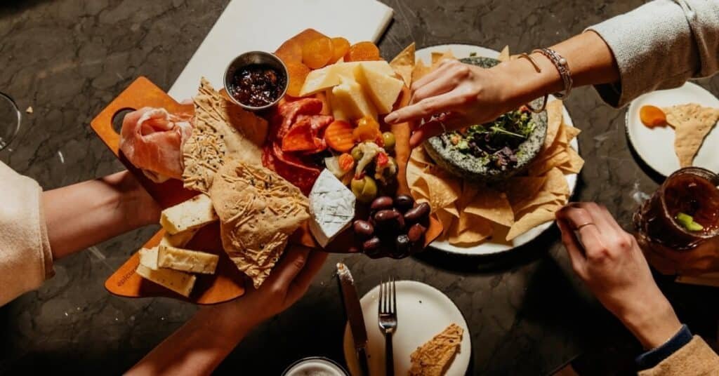 Hands sharing a large Tiger Brunch charcuterie and cheese board with crackers, fruit, olives, and dip