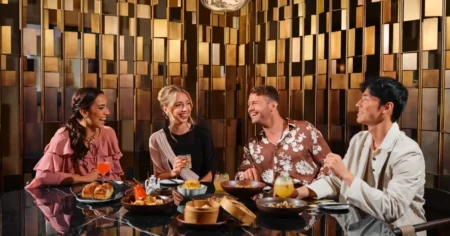 Four friends laugh over drinks and dishes at a restaurant table with a gold geometric wall behind them