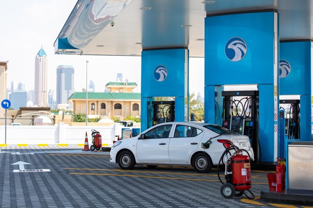 UAE Petrol Prices May 2026: A white car at a gas station with blue and white pumps, urban buildings visible in the background.