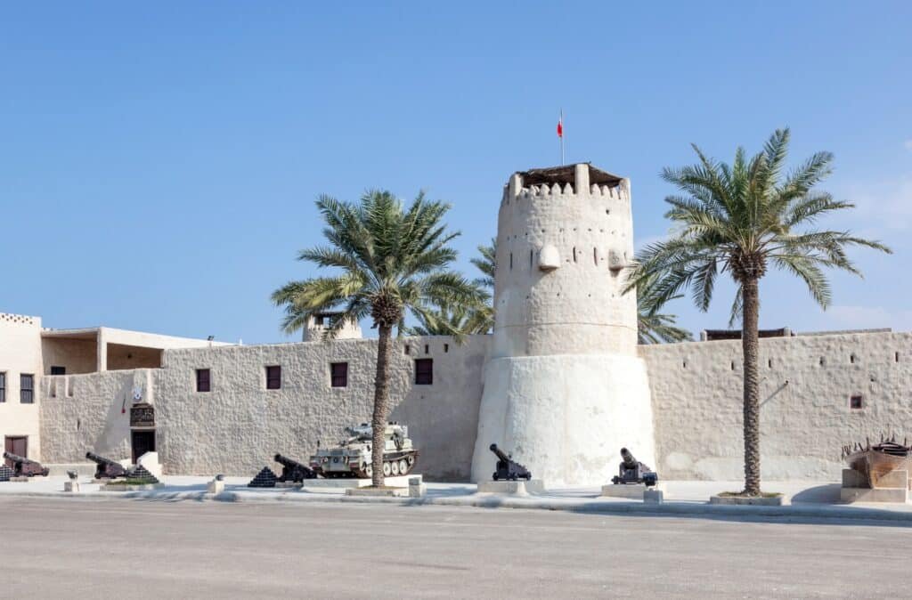 Desert fort with a white tower, palm trees, and mounted cannons under a clear blue sky