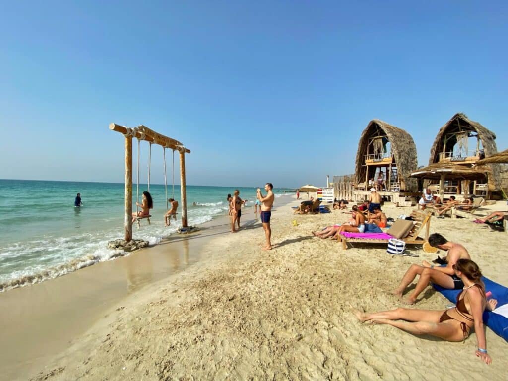 Sunny beach scene with people lounging, swimming, and riding a wooden swing beside thatched cabanas by the sea