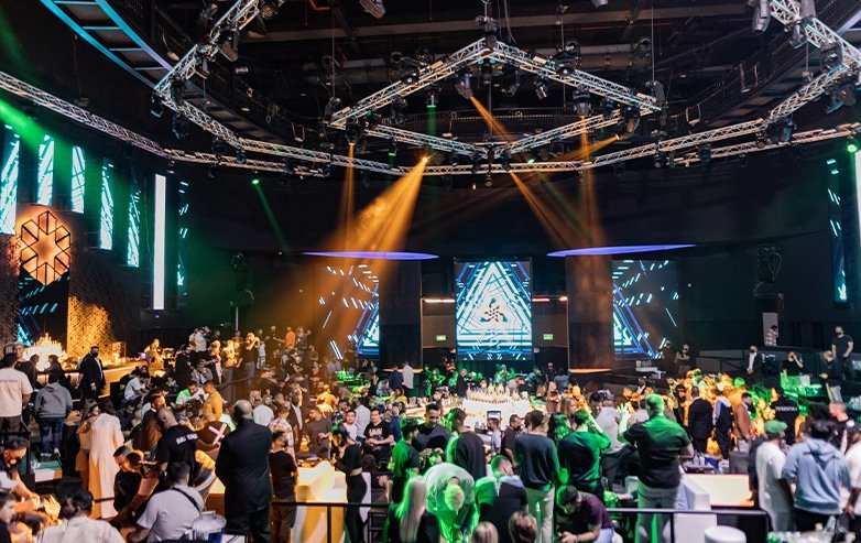Crowded event venue with a large stage, bright screens, and green and orange spotlights over attendees