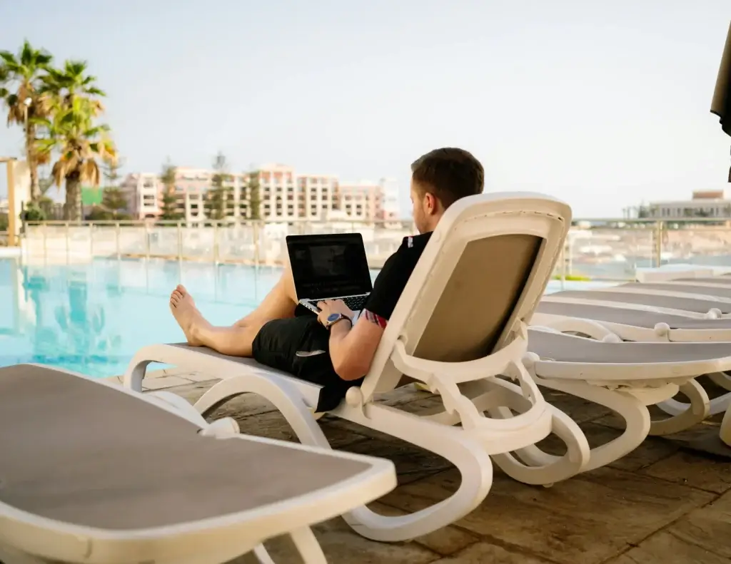 Workcations In The UAE: A person lounging on a deck chair by a pool, working on a laptop.