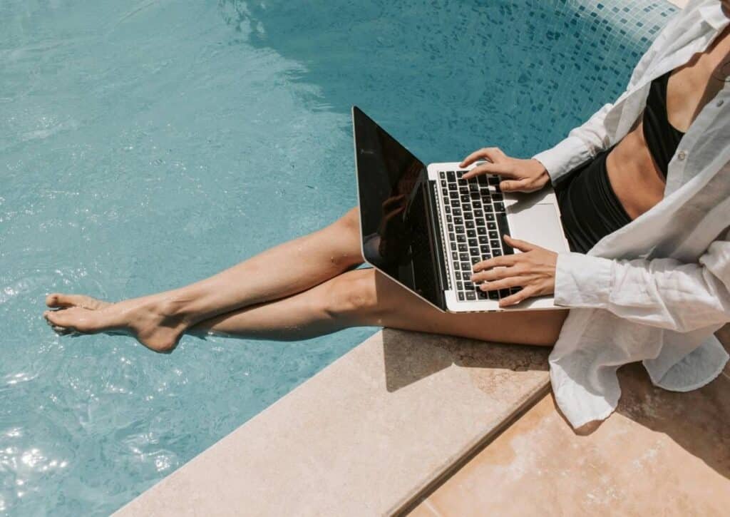 Person in casual attire sitting by a pool, using a laptop with their legs dipped in the water.