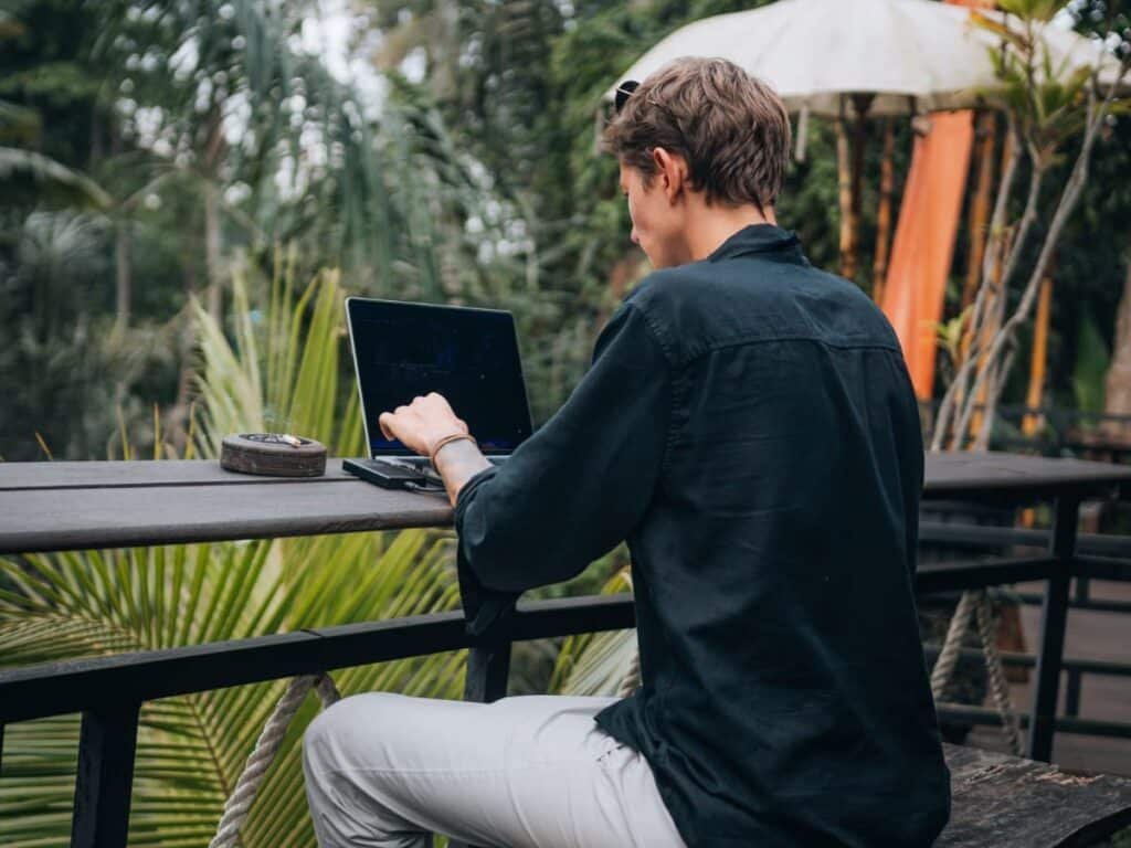 Person sitting outdoors at a laptop on a wooden table, surrounded by tropical plants.