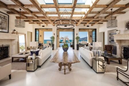 Luxurious living room with wooden ceiling beams, a long wooden table, sofas, chandeliers, and large windows revealing a scenic view.