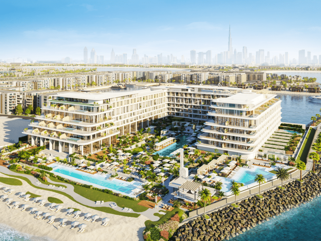 Aerial view of luxury beachfront resort with two modern buildings, pools, and landscaped gardens, city skyline in the background.