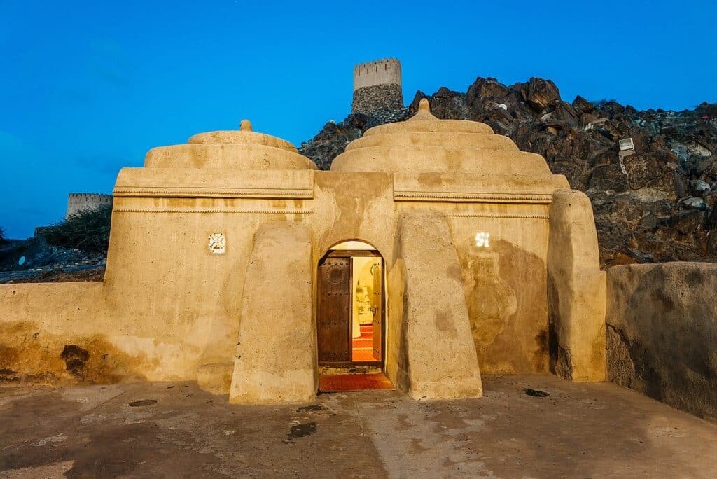 Mud fort entrance at dusk beneath a rocky hill with watchtowers and a bright blue sky