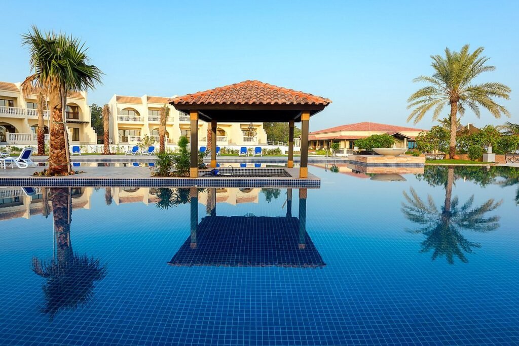 Resort pool with a tiled gazebo, palm trees, and villas reflected in the still water