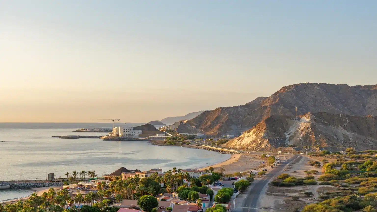 Sunset over a calm coastal bay with a beach, palm trees, roads, and rocky mountains along the shoreline