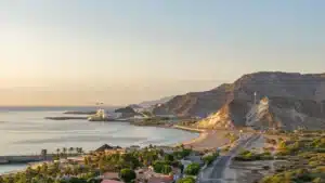 Sunset over a calm coastal bay with a beach, palm trees, roads, and rocky mountains along the shoreline