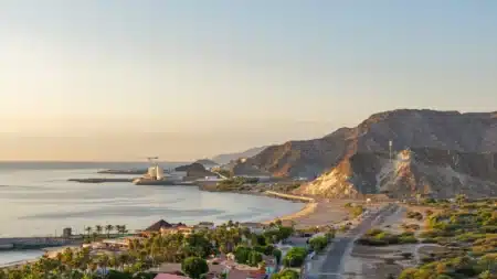 Sunset over a calm coastal bay with a beach, palm trees, roads, and rocky mountains along the shoreline