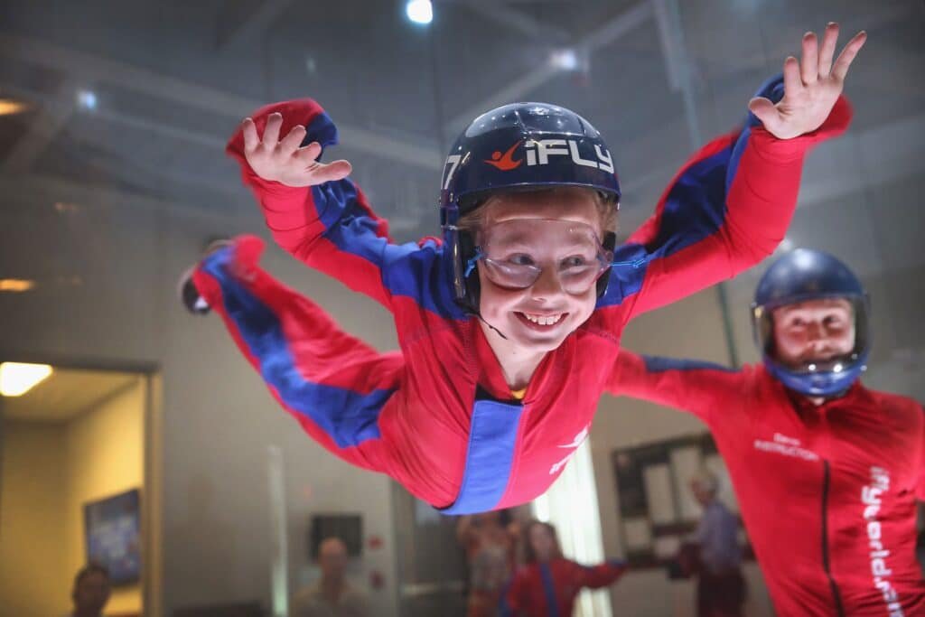 Smiling child in a red and blue jumpsuit skydiving in an indoor wind tunnel with an instructor behind them