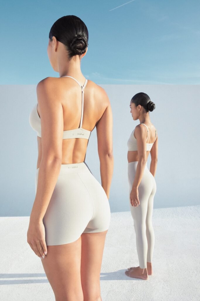 Two women in beige activewear stand on a white surface against a blue sky, one facing forward and the other sideways.