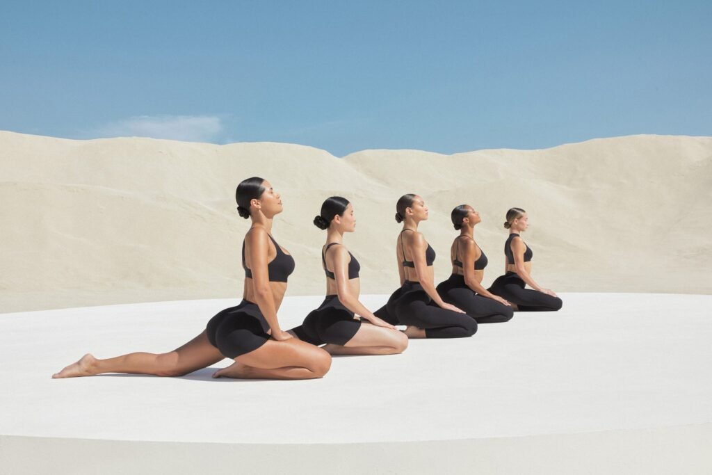Five women in black NikeSKIMS Studio Stretch athletic wear seated in a kneeling pose on a white circular platform against a sandy landscape and blue sky.
