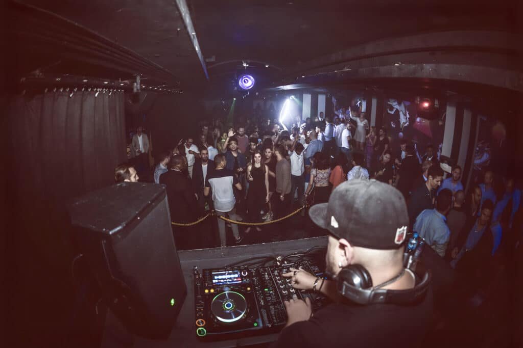 Smokingroove mixing music in a crowded nightclub dance floor with colorful lights and raised hands