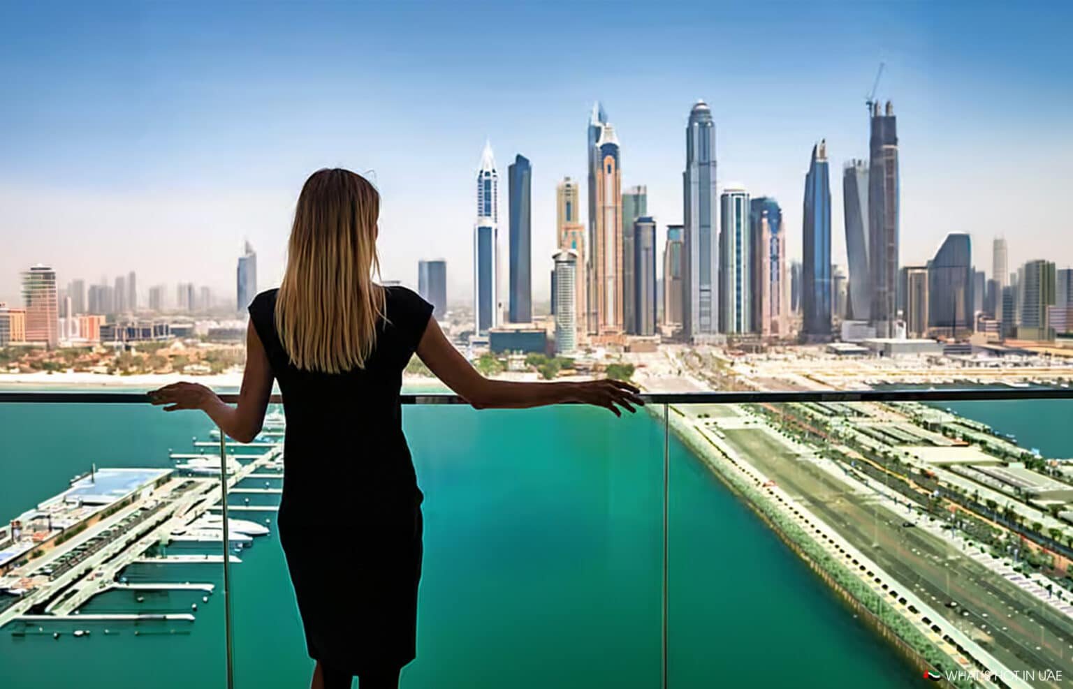 Woman in a black dress stands on a balcony overlooking a waterfront skyline of tall skyscrapers