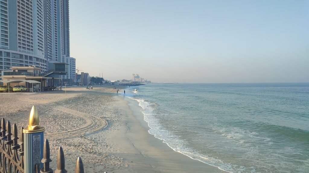 Sandy beach beside tall waterfront towers, with calm sea and a few people near the shore at sunset