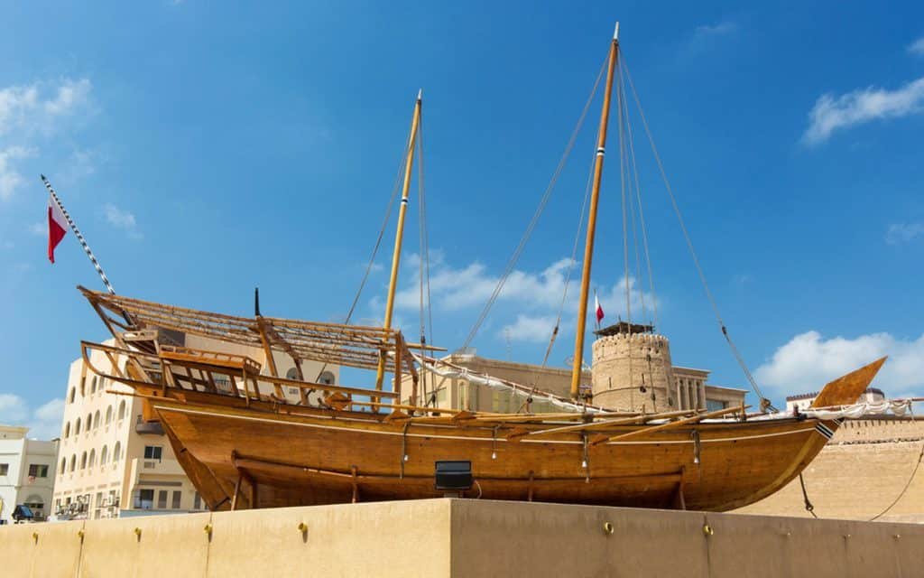 Large wooden sailing dhow displayed on a pedestal beside historic buildings under a blue sky