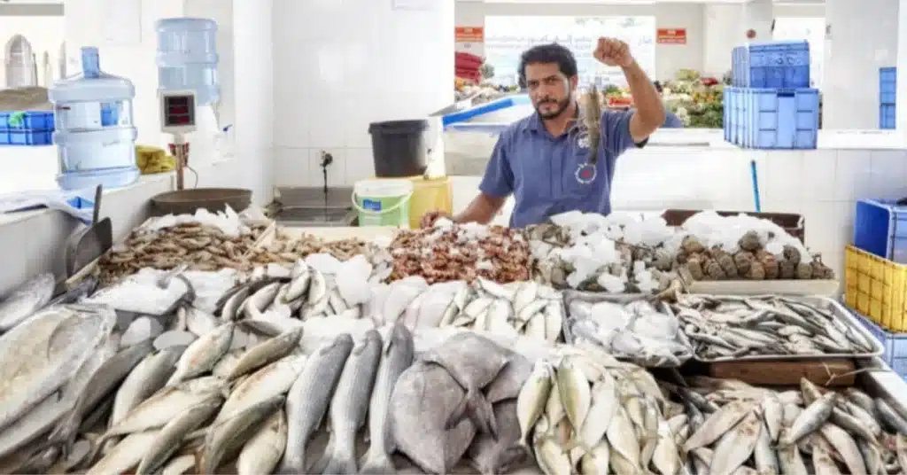 Fish vendor holding up a small fish behind a crowded seafood display in a market stall