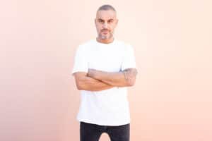 Man with short gray hair and a tattooed arm, wearing a white T-shirt and black jeans, standing with arms crossed against a pink wall