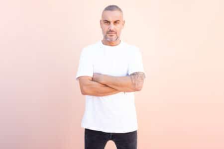 Man with short gray hair and a tattooed arm, wearing a white T-shirt and black jeans, standing with arms crossed against a pink wall